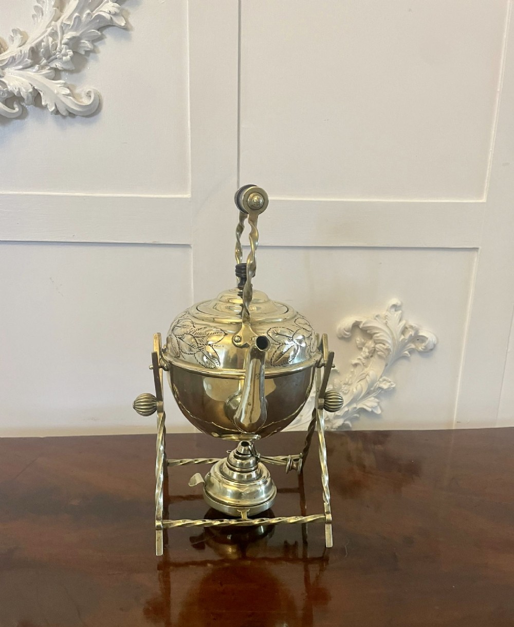 An antique Victorian brass spirit kettle featuring floral engravings, an ebonised wooden handle, and a matching brass stand with a spirit burner. View X