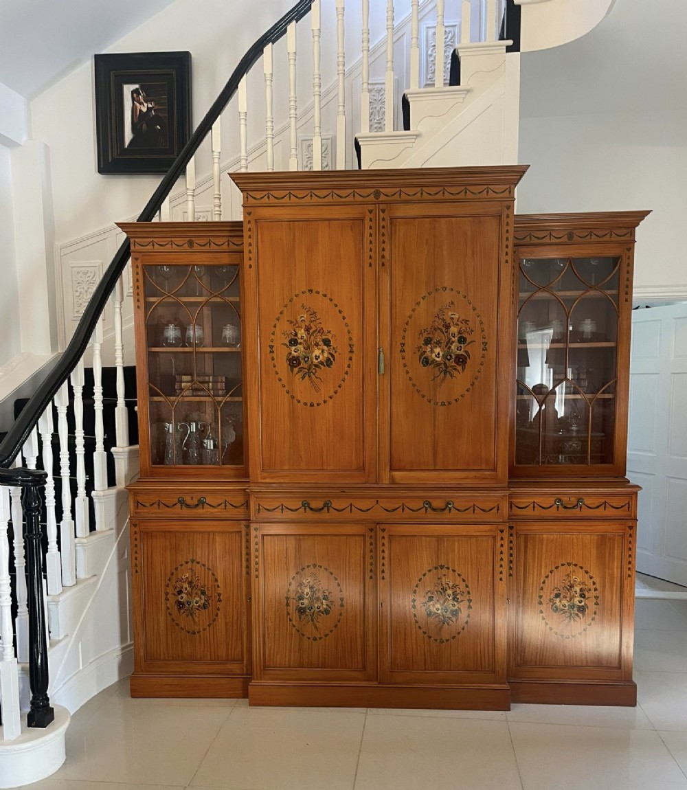 A large satinwood breakfront bookcase featuring hand-painted floral cartouches, upper glazed display doors, and lower cupboard panels. View II