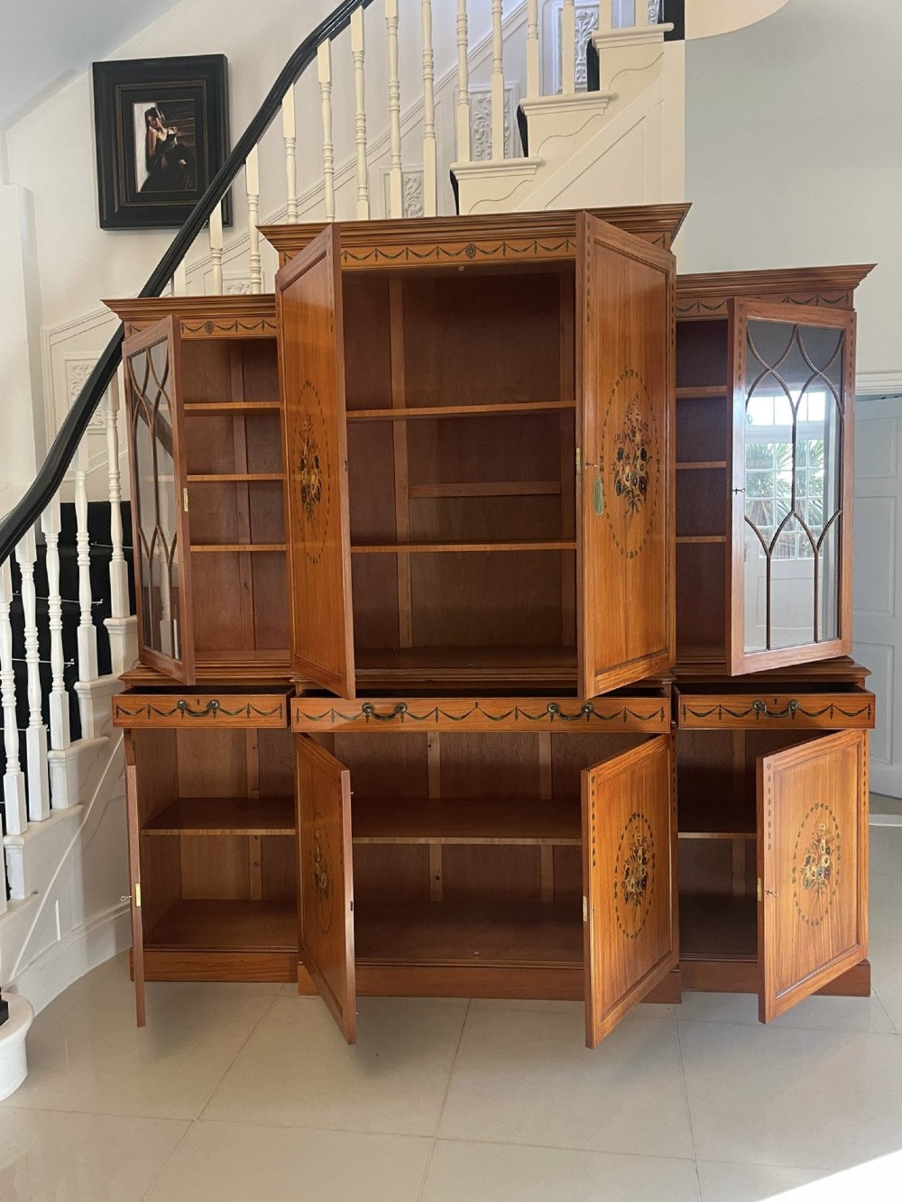 A large satinwood breakfront bookcase featuring hand-painted floral cartouches, upper glazed display doors, and lower cupboard panels. View III