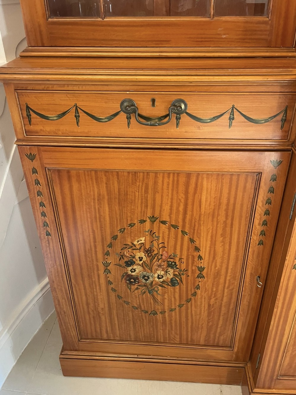 A large satinwood breakfront bookcase featuring hand-painted floral cartouches, upper glazed display doors, and lower cupboard panels. View IV