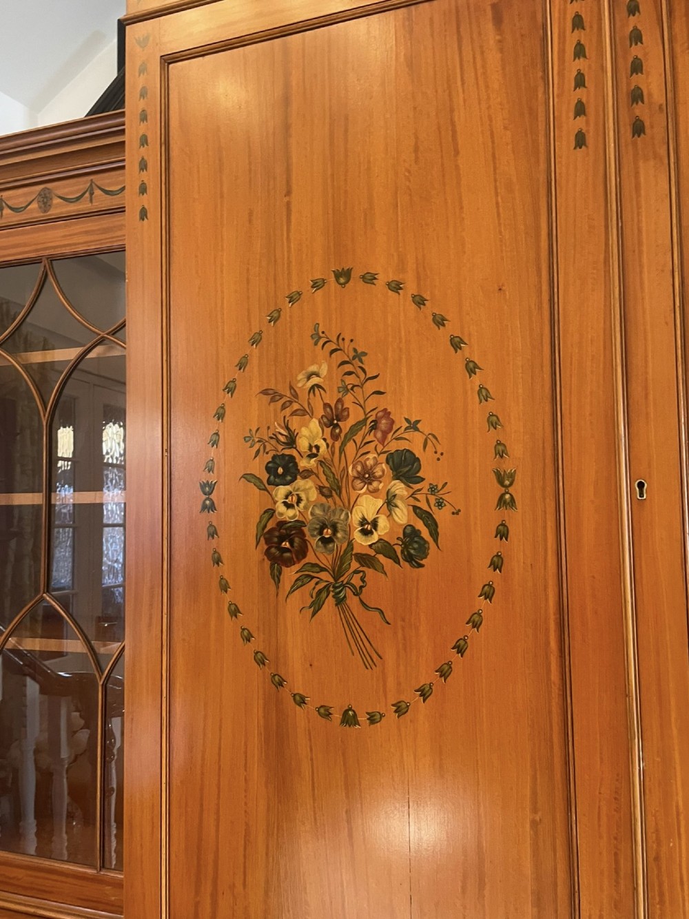 A large satinwood breakfront bookcase featuring hand-painted floral cartouches, upper glazed display doors, and lower cupboard panels. View V