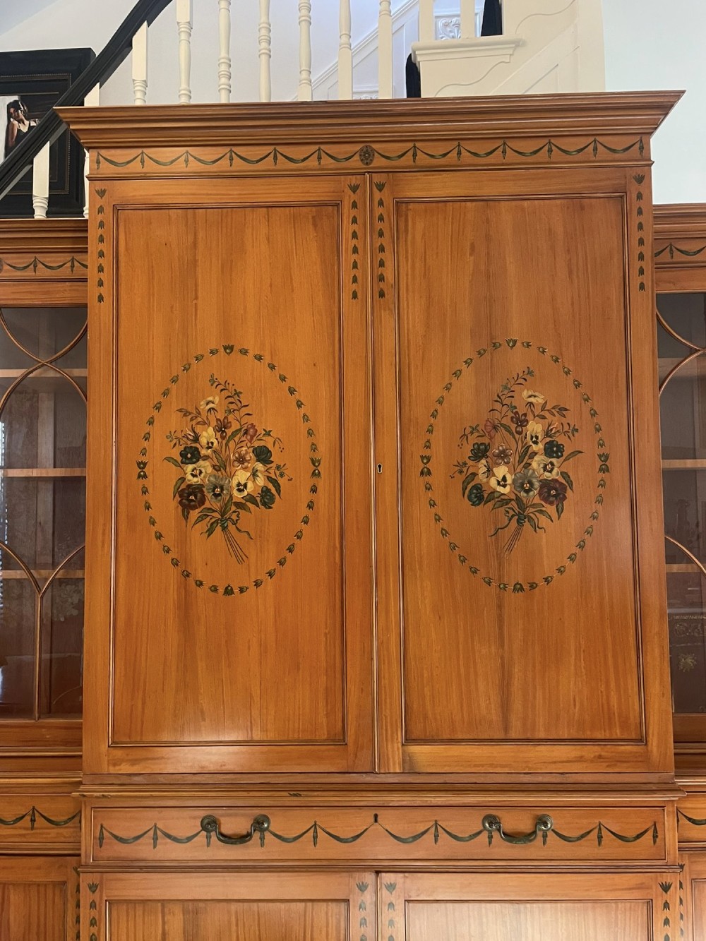 A large satinwood breakfront bookcase featuring hand-painted floral cartouches, upper glazed display doors, and lower cupboard panels. View VI