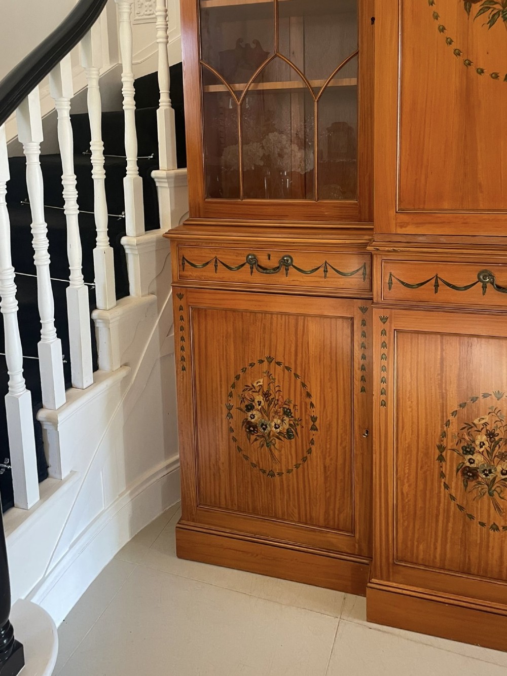A large satinwood breakfront bookcase featuring hand-painted floral cartouches, upper glazed display doors, and lower cupboard panels. View VII