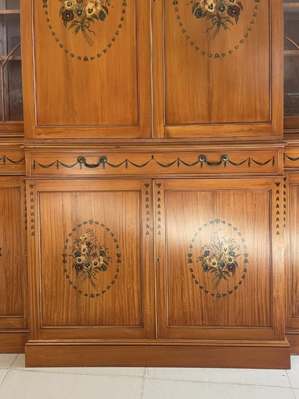 A large satinwood breakfront bookcase featuring hand-painted floral cartouches, upper glazed display doors, and lower cupboard panels. View VIII