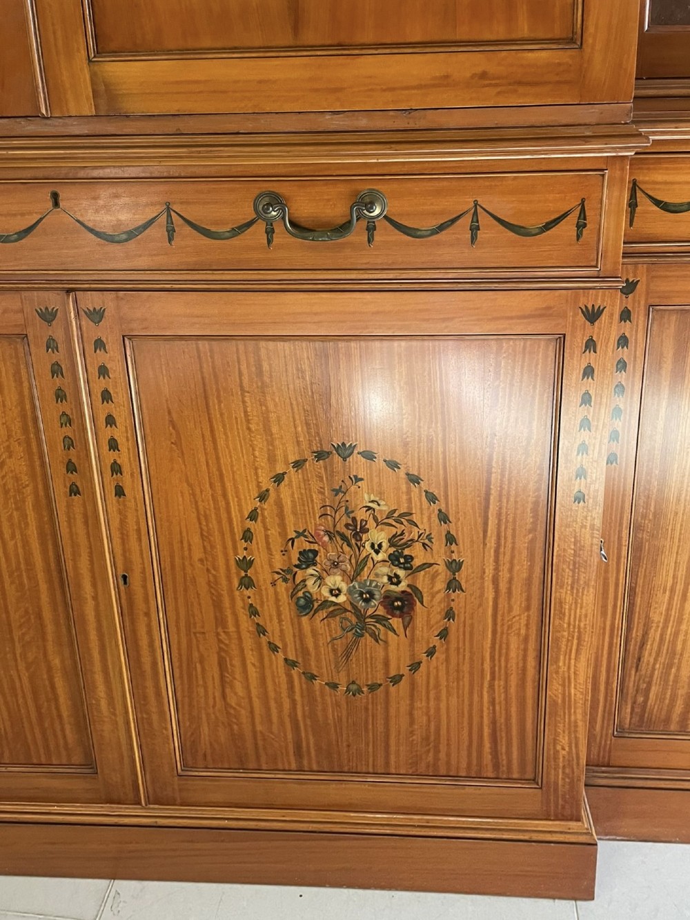 A large satinwood breakfront bookcase featuring hand-painted floral cartouches, upper glazed display doors, and lower cupboard panels. View X