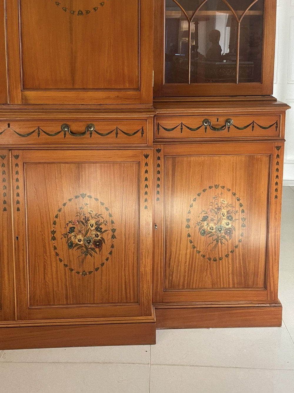A large satinwood breakfront bookcase featuring hand-painted floral cartouches, upper glazed display doors, and lower cupboard panels. View XII