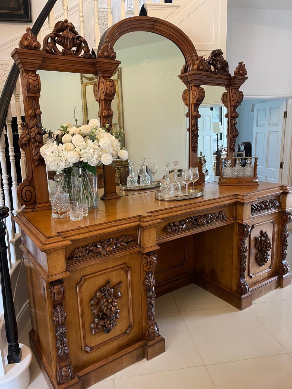 A large Victorian carved oak sideboard featuring three upper mirror panels, intricate grape cluster wood carvings, and a pedestal base. View V