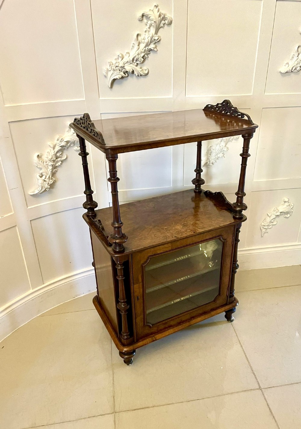 An antique Victorian burr walnut music cabinet featuring two open upper shelves supported by turned columns and a lower glass-fronted cupboard with gold lettered divisions on castors. View II