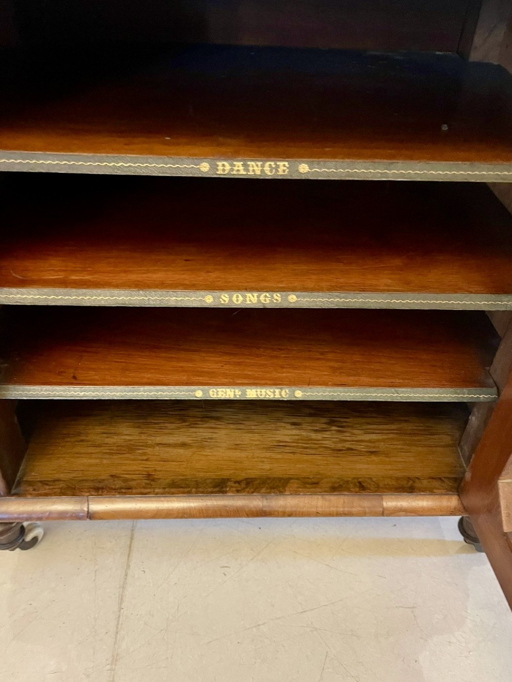 An antique Victorian burr walnut music cabinet featuring two open upper shelves supported by turned columns and a lower glass-fronted cupboard with gold lettered divisions on castors. View IV