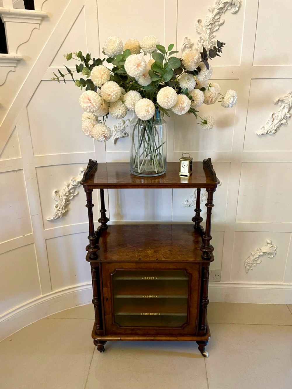 An antique Victorian burr walnut music cabinet featuring two open upper shelves supported by turned columns and a lower glass-fronted cupboard with gold lettered divisions on castors. View VI