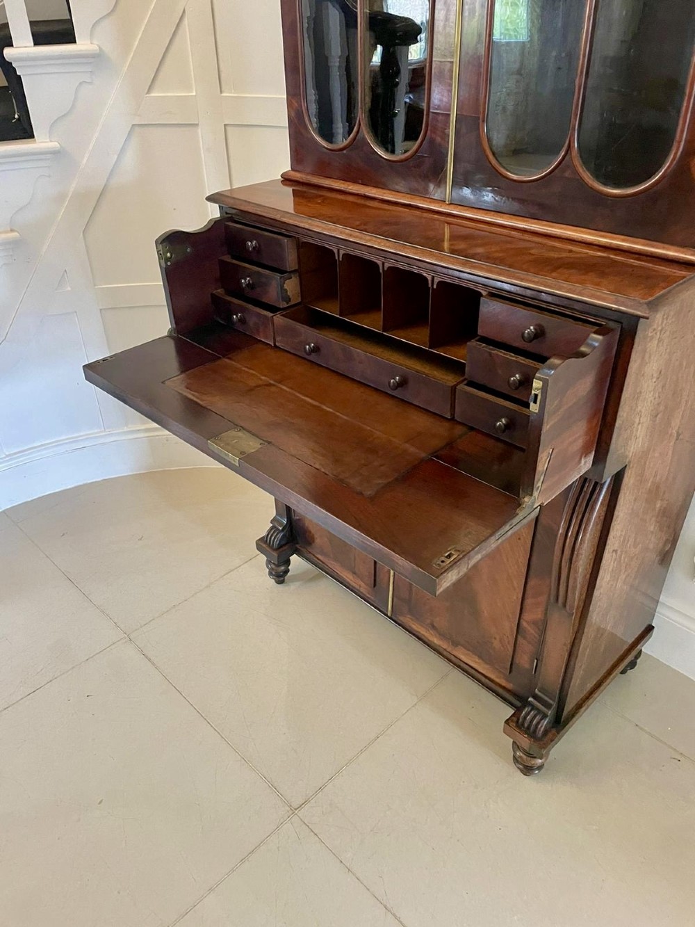 An antique Regency mahogany secretaire bookcase featuring a carved scroll pediment, glazed upper doors, a fall-front desk drawer, and a lower two-door cupboard. View III