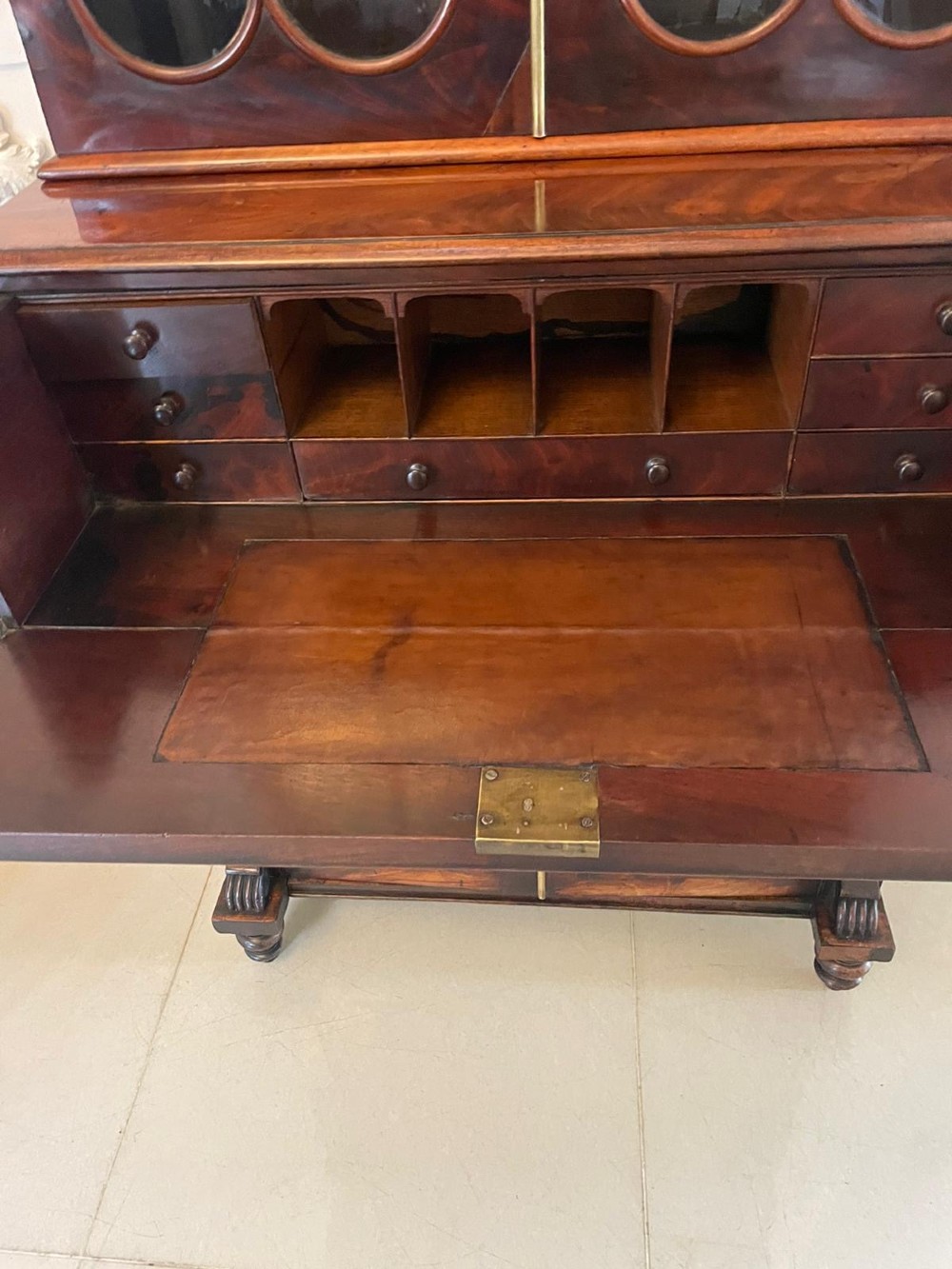 An antique Regency mahogany secretaire bookcase featuring a carved scroll pediment, glazed upper doors, a fall-front desk drawer, and a lower two-door cupboard. View IV