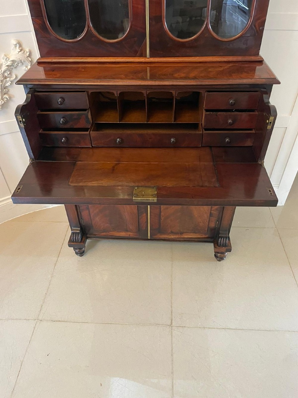 An antique Regency mahogany secretaire bookcase featuring a carved scroll pediment, glazed upper doors, a fall-front desk drawer, and a lower two-door cupboard. View V