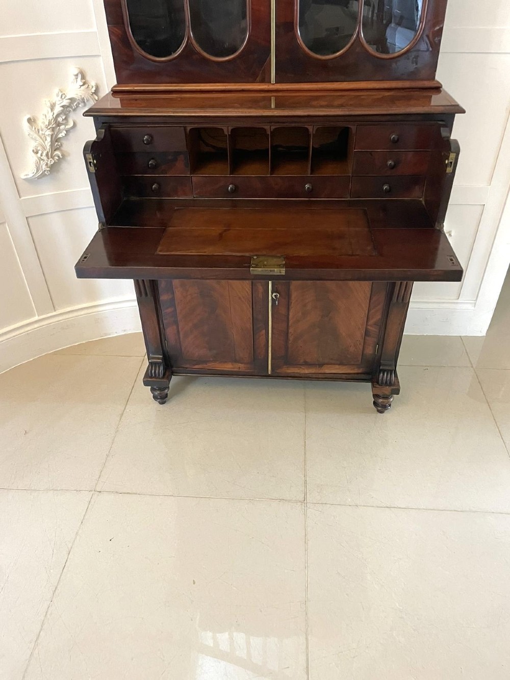 An antique Regency mahogany secretaire bookcase featuring a carved scroll pediment, glazed upper doors, a fall-front desk drawer, and a lower two-door cupboard. View VI