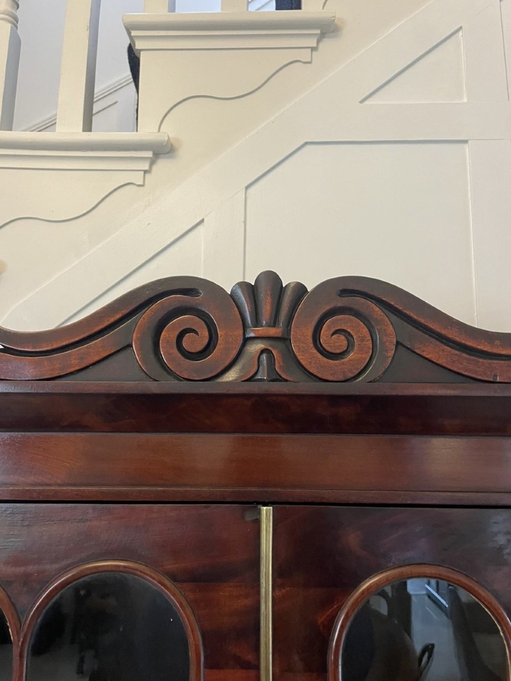 An antique Regency mahogany secretaire bookcase featuring a carved scroll pediment, glazed upper doors, a fall-front desk drawer, and a lower two-door cupboard. View IX