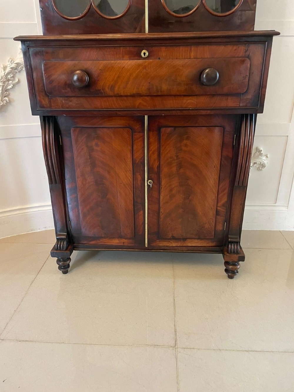 An antique Regency mahogany secretaire bookcase featuring a carved scroll pediment, glazed upper doors, a fall-front desk drawer, and a lower two-door cupboard. View XIV