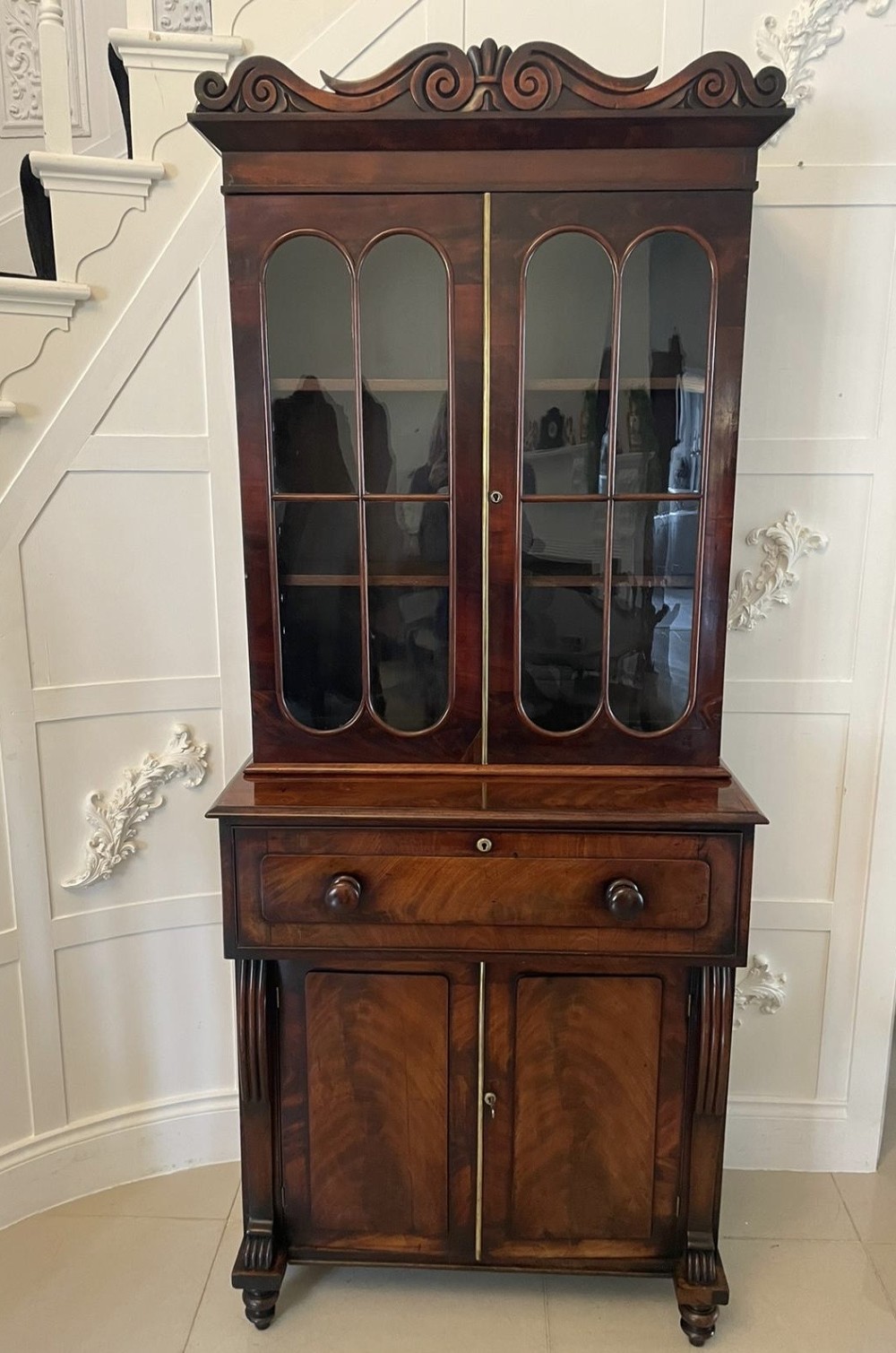 An antique Regency mahogany secretaire bookcase featuring a carved scroll pediment, glazed upper doors, a fall-front desk drawer, and a lower two-door cupboard.