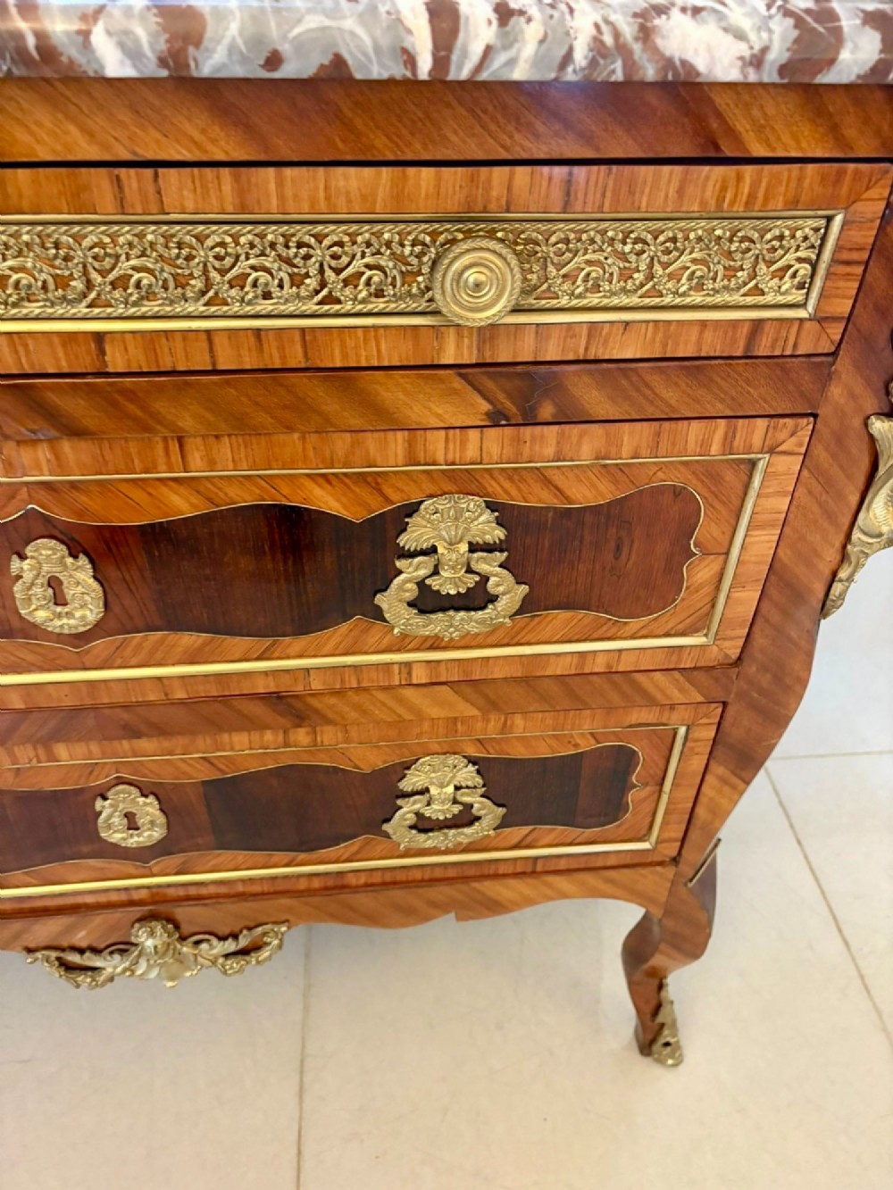 An antique French style commode chest featuring a dark grey veined marble top, decorative walnut veneers, cast brass mounts, and cabriole legs. View V