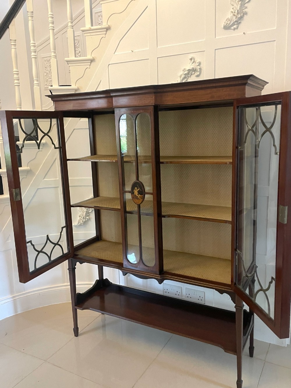 An Edwardian mahogany display cabinet featuring glazed doors with astragal bars, a central marquetry inlay medallion, tapering square legs, and an undertier shelf. View II
