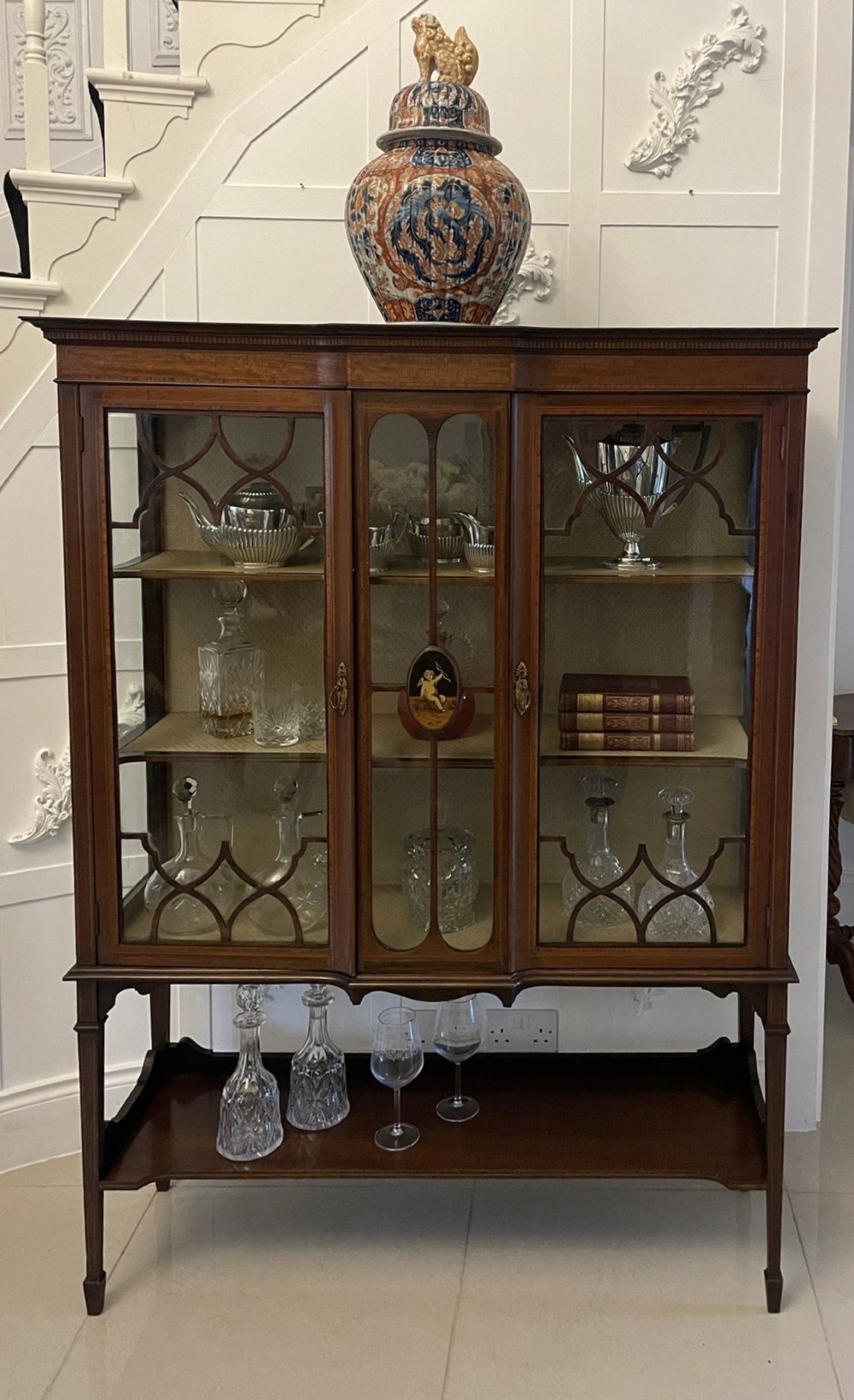 An Edwardian mahogany display cabinet featuring glazed doors with astragal bars, a central marquetry inlay medallion, tapering square legs, and an undertier shelf. View V