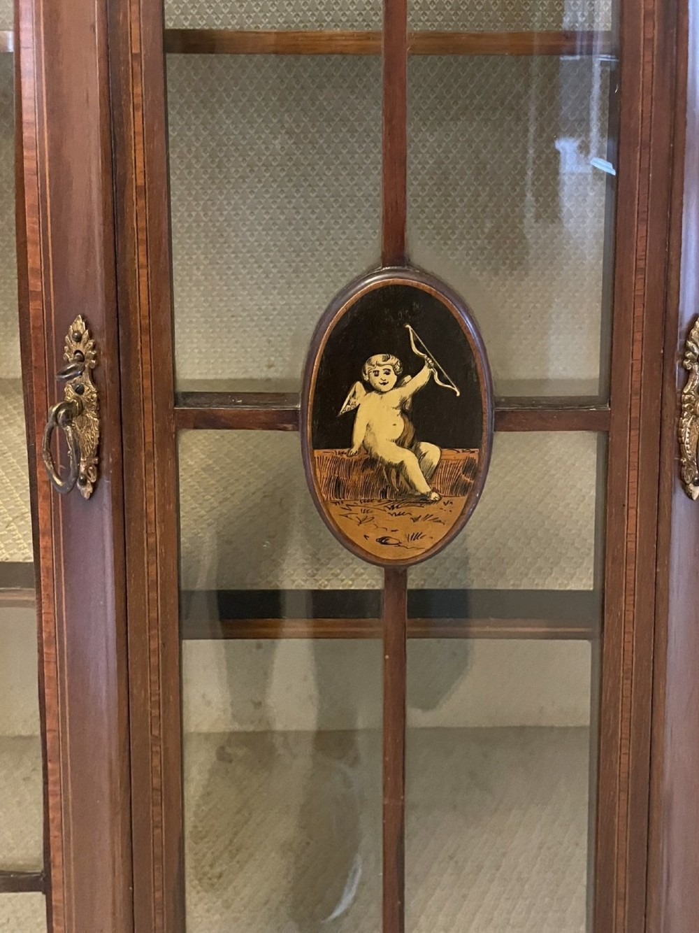 An Edwardian mahogany display cabinet featuring glazed doors with astragal bars, a central marquetry inlay medallion, tapering square legs, and an undertier shelf. View VI
