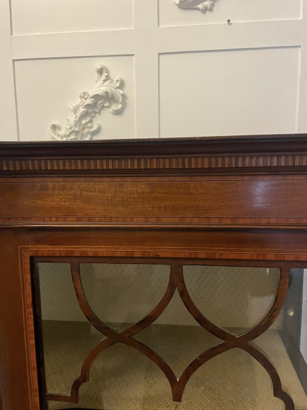 An Edwardian mahogany display cabinet featuring glazed doors with astragal bars, a central marquetry inlay medallion, tapering square legs, and an undertier shelf. View IX