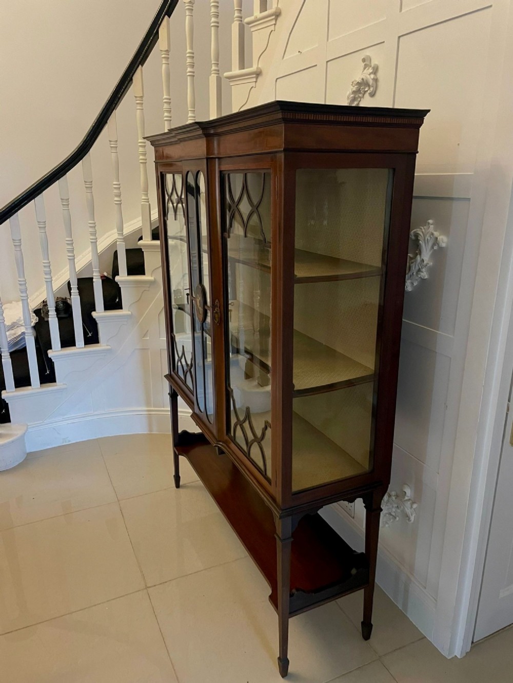 An Edwardian mahogany display cabinet featuring glazed doors with astragal bars, a central marquetry inlay medallion, tapering square legs, and an undertier shelf. View X