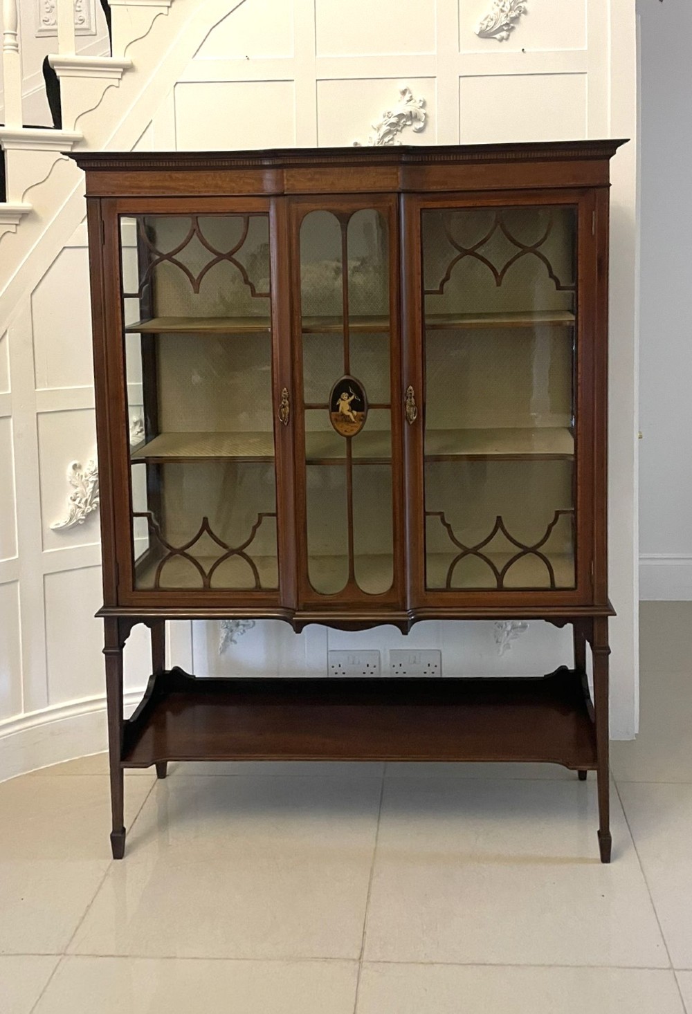 An Edwardian mahogany display cabinet featuring glazed doors with astragal bars, a central marquetry inlay medallion, tapering square legs, and an undertier shelf.