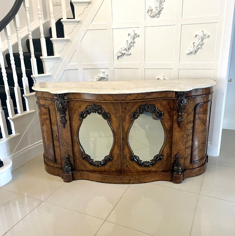 A Victorian burr walnut credenza featuring a serpentine white marble top above two central oval mirrored doors and carved scroll accents.