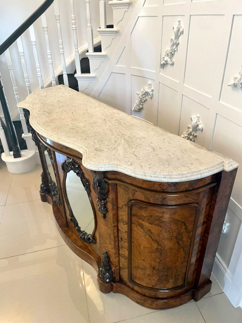 A Victorian burr walnut credenza featuring a serpentine white marble top above two central oval mirrored doors and carved scroll accents. View IV