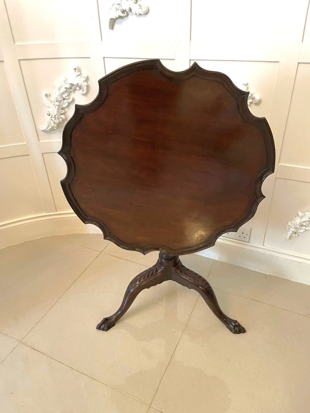 An antique George III mahogany tilt-top table featuring a scalloped pie-crust rim supported by a carved central pedestal and tripod cabriole legs.