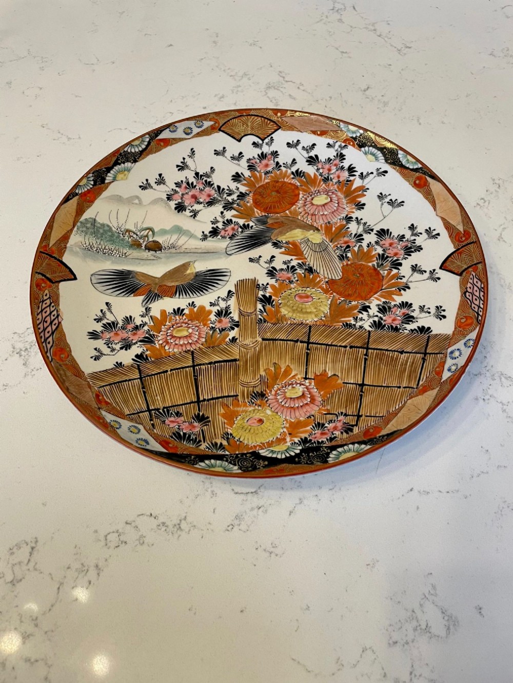 A shallow circular Japanese Kutani porcelain bowl featuring hand-painted floral chrysanthemum patterns, birds in flight, and detailed decorative borders in terracotta, red, black, and gold.