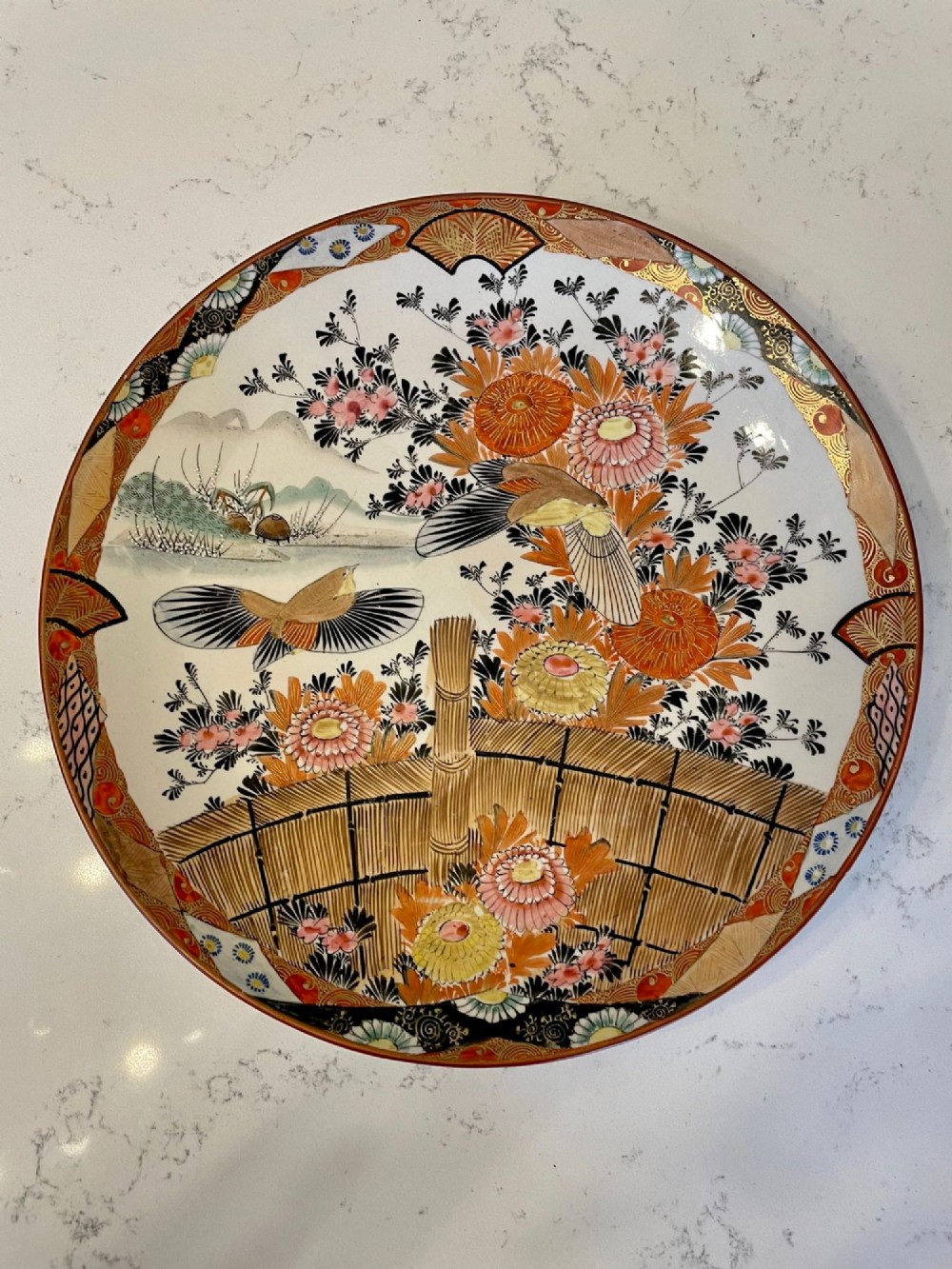 A shallow circular Japanese Kutani porcelain bowl featuring hand-painted floral chrysanthemum patterns, birds in flight, and detailed decorative borders in terracotta, red, black, and gold. View VII