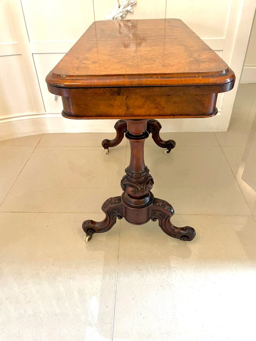 A Victorian burr walnut fold-over card table featuring a rectangular figured veneer top with rounded corners, twin turned pedestals, and carved legs on castors. View VII