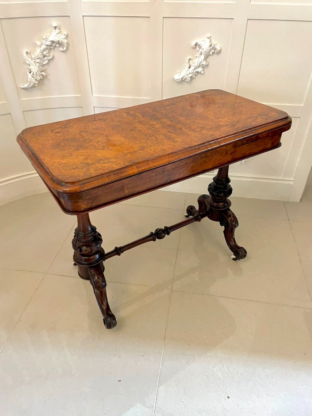 A Victorian burr walnut fold-over card table featuring a rectangular figured veneer top with rounded corners, twin turned pedestals, and carved legs on castors.