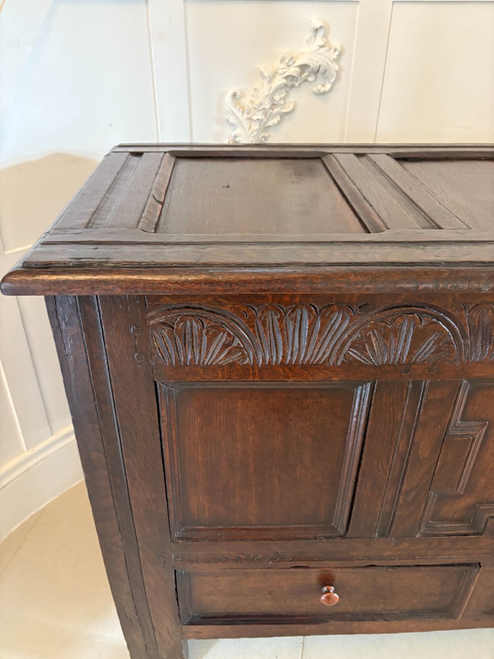 A 17th-century dark oak mule chest featuring a hinged paneled lid, a carved frieze above geometric front panels, and two lower drawers with wooden knobs. View XII