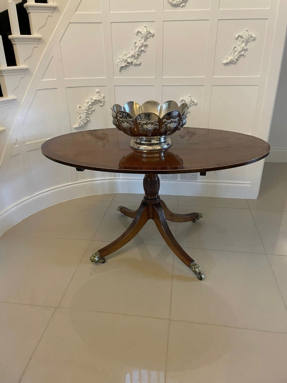 An antique George III oval tilt-top dining table in flame mahogany, featuring a crossbanded border and a quadruped pedestal base with reeded legs and brass castors. View II