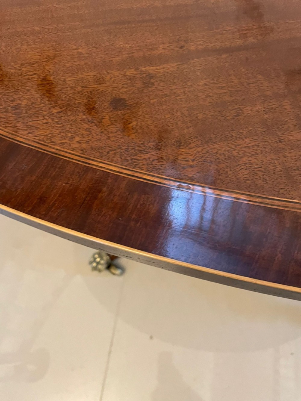 An antique George III oval tilt-top dining table in flame mahogany, featuring a crossbanded border and a quadruped pedestal base with reeded legs and brass castors. View III