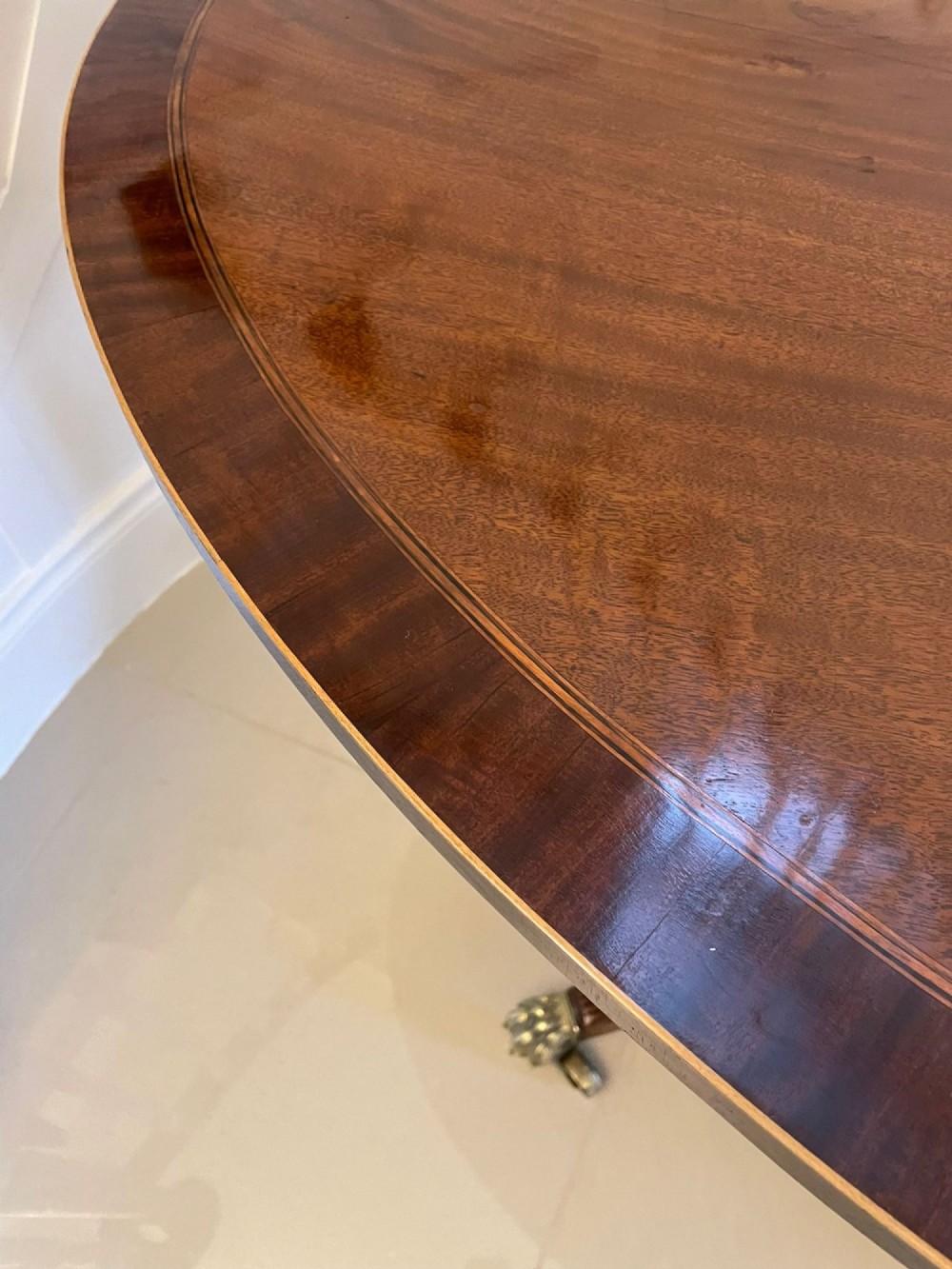 An antique George III oval tilt-top dining table in flame mahogany, featuring a crossbanded border and a quadruped pedestal base with reeded legs and brass castors. View IV