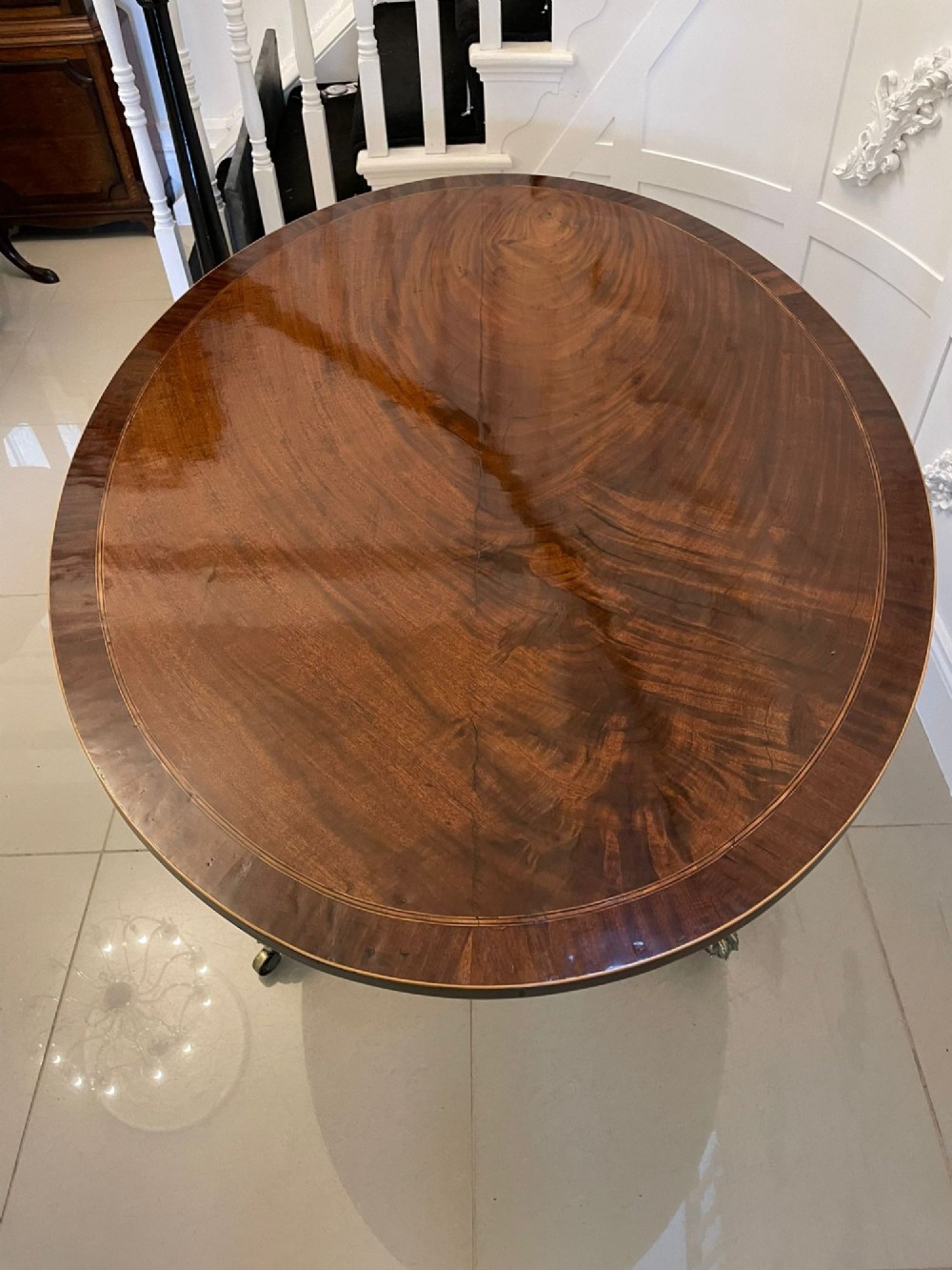 An antique George III oval tilt-top dining table in flame mahogany, featuring a crossbanded border and a quadruped pedestal base with reeded legs and brass castors. View V