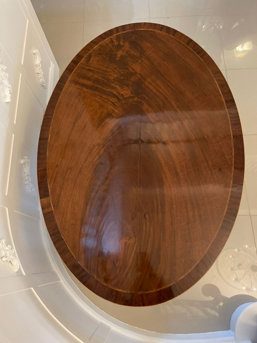 An antique George III oval tilt-top dining table in flame mahogany, featuring a crossbanded border and a quadruped pedestal base with reeded legs and brass castors. View VI