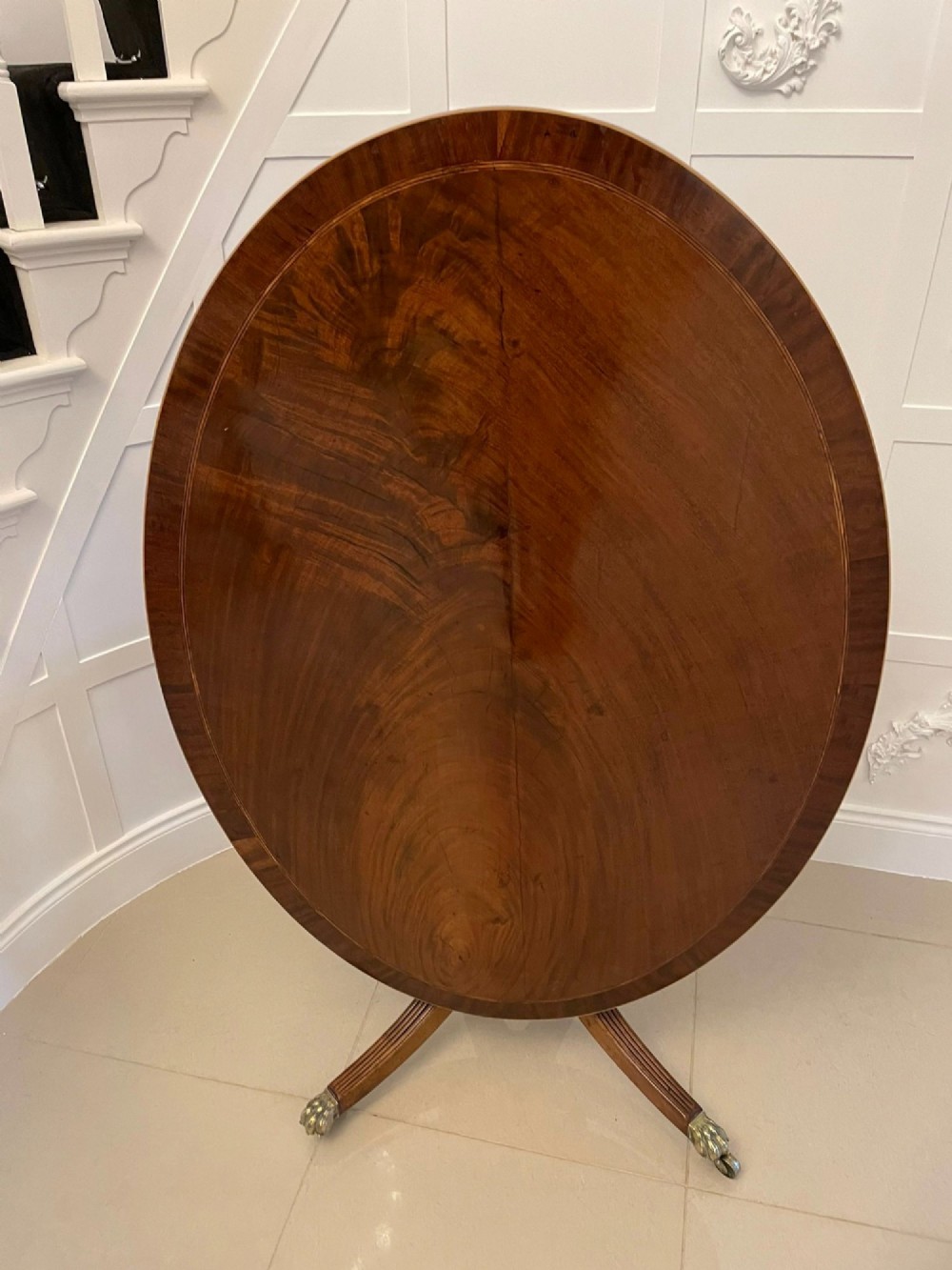 An antique George III oval tilt-top dining table in flame mahogany, featuring a crossbanded border and a quadruped pedestal base with reeded legs and brass castors.