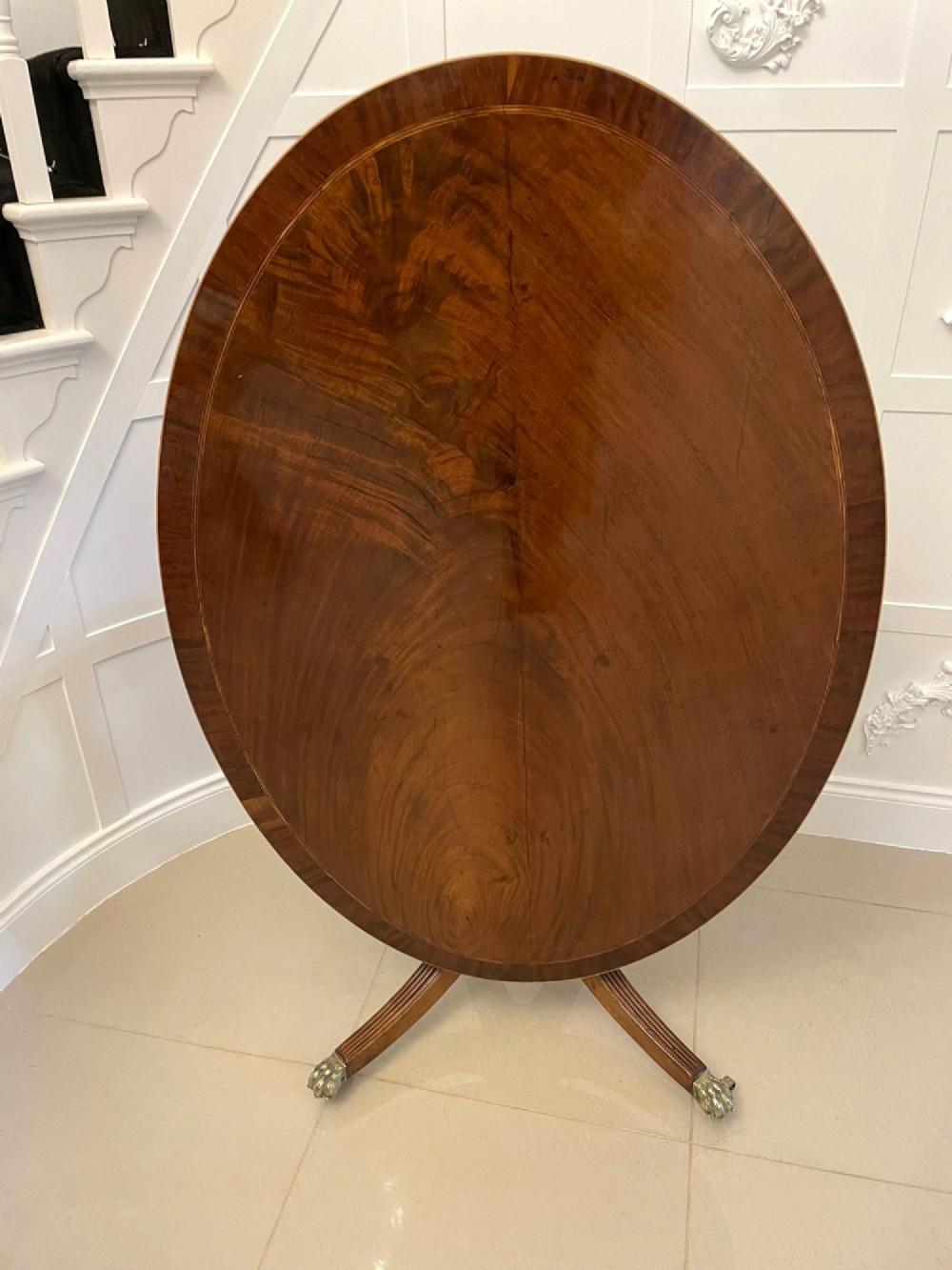 An antique George III oval tilt-top dining table in flame mahogany, featuring a crossbanded border and a quadruped pedestal base with reeded legs and brass castors. View VII