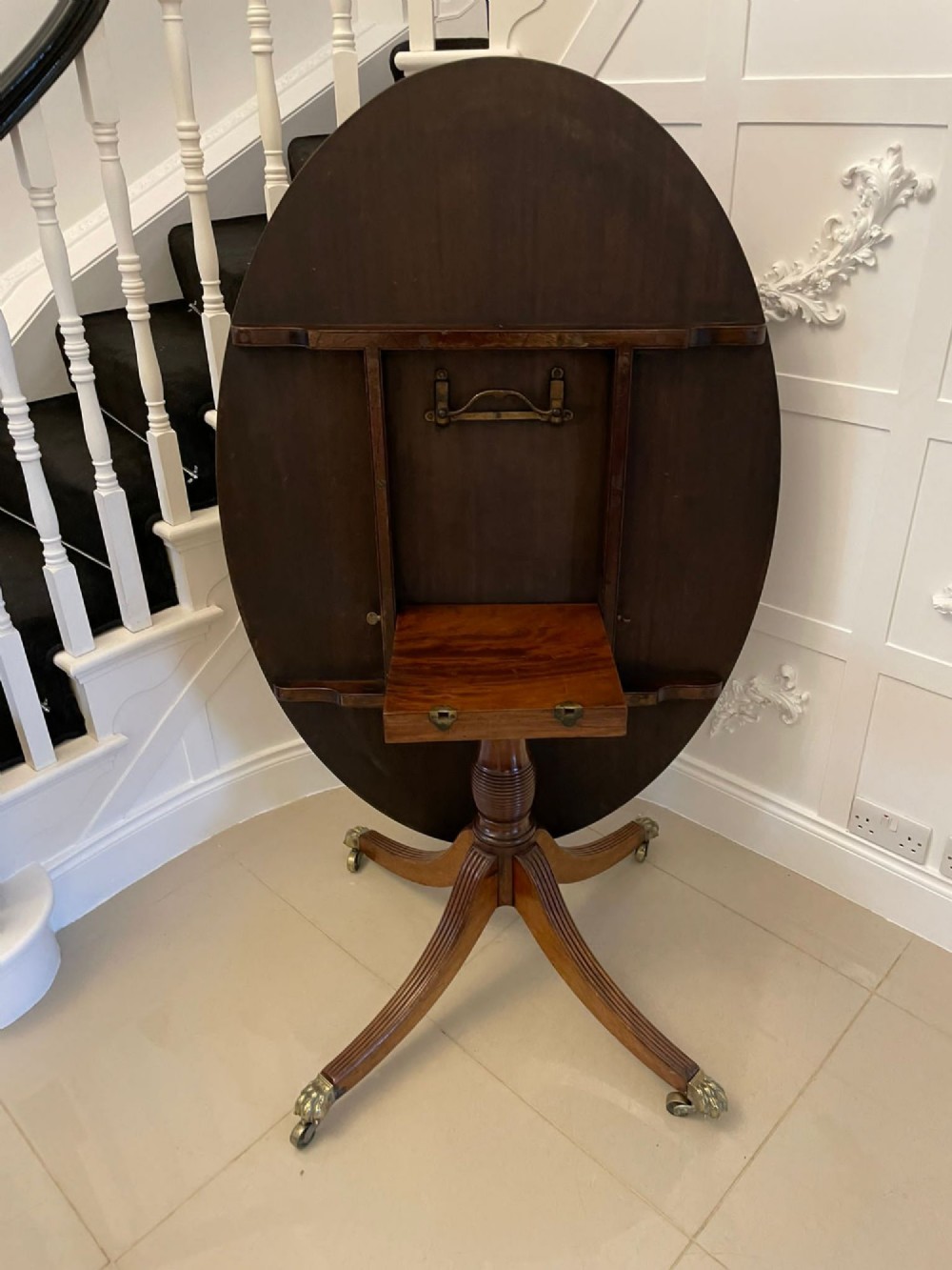 An antique George III oval tilt-top dining table in flame mahogany, featuring a crossbanded border and a quadruped pedestal base with reeded legs and brass castors. View VIII