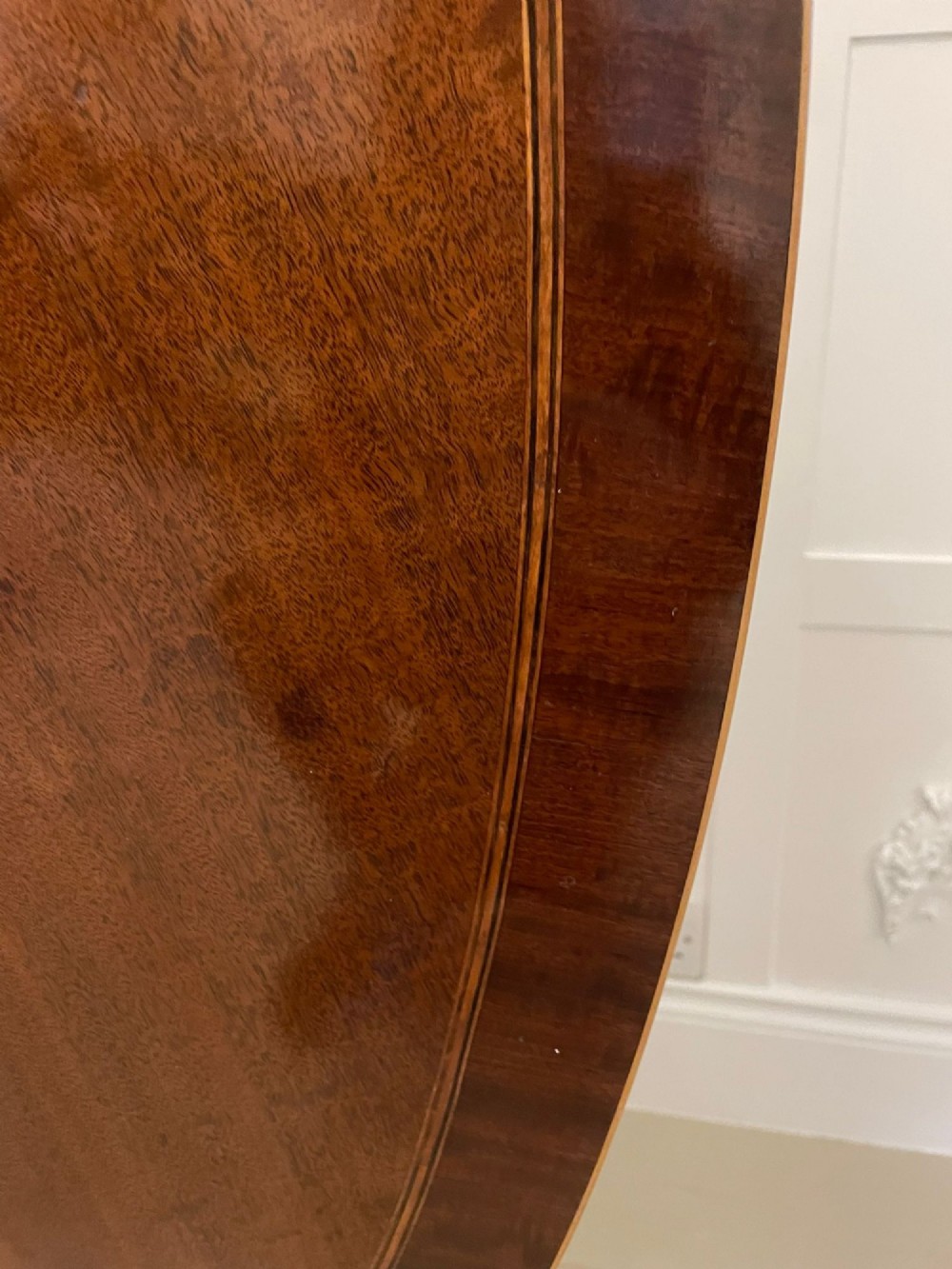 An antique George III oval tilt-top dining table in flame mahogany, featuring a crossbanded border and a quadruped pedestal base with reeded legs and brass castors. View X