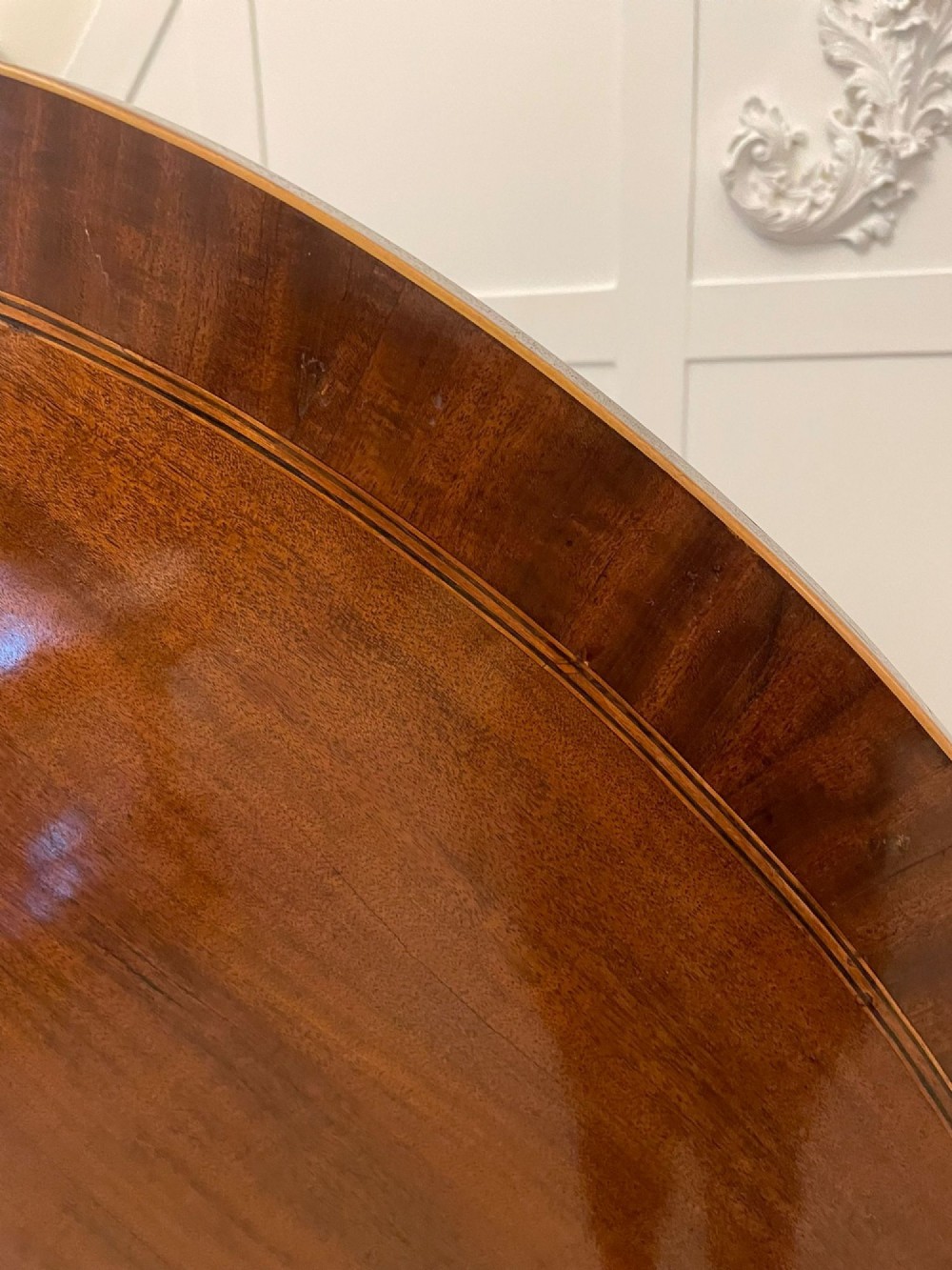 An antique George III oval tilt-top dining table in flame mahogany, featuring a crossbanded border and a quadruped pedestal base with reeded legs and brass castors. View XII