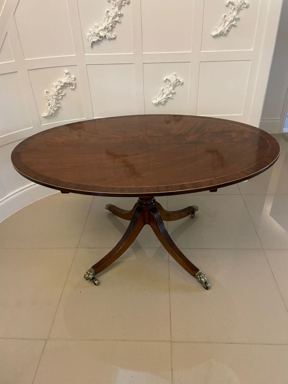 An antique George III oval tilt-top dining table in flame mahogany, featuring a crossbanded border and a quadruped pedestal base with reeded legs and brass castors. View XV