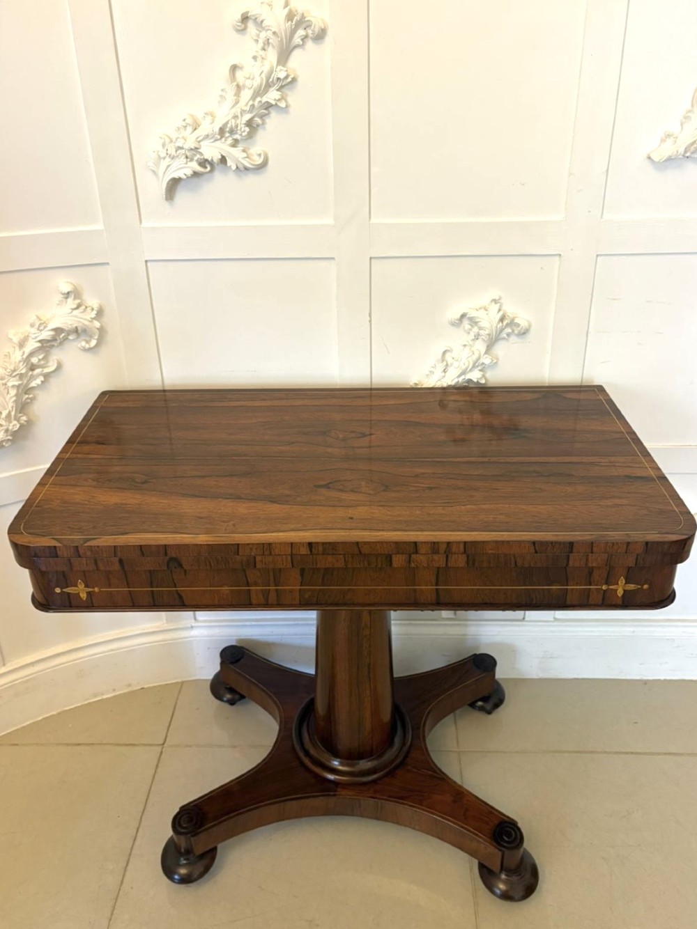 A Regency rosewood card table featuring fine brass inlay stringing, a fold-over swivel top, a central pedestal stem, and a quadform base on round feet. View III