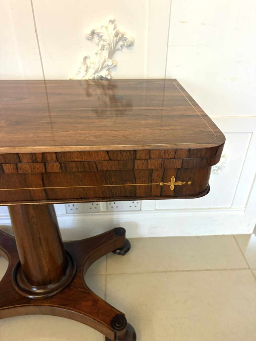 A Regency rosewood card table featuring fine brass inlay stringing, a fold-over swivel top, a central pedestal stem, and a quadform base on round feet. View IV