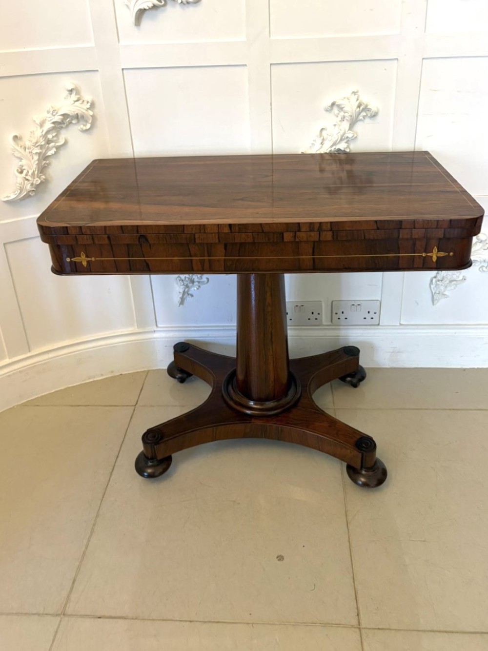 A Regency rosewood card table featuring fine brass inlay stringing, a fold-over swivel top, a central pedestal stem, and a quadform base on round feet.